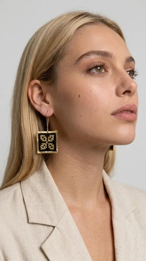 The embroidered statement earrings with geometric patterns in black are the ideal choice for those seeking a piece of jewelry that combines folk tradition with modern design. Designed by Greek Jewelry Designer Aikaterini Chalkiadaki, these handmade earrings are a unique piece that stands out instantly thanks to their square shape and striking embroidery with golden details on black fabric.

Made from brass and fabric, these earrings feature a geometric motif inspired by folklore aesthetics — four symmetrical embroidered elements in a gold tone are framed by a golden brass frame, creating a result reminiscent of traditional Greek handicrafts. They hang from a gold hook and add movement and character to any look.

Ideal for weddings, christenings, evening outings, or even to elevate a casual everyday look, these statement earrings pair beautifully with both white outfits and neutral colors, as shown in the photo.