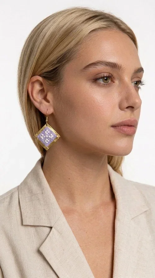 These embroidered statement earrings in a lilac shade are the ideal choice for those seeking an accessory that combines traditional craftsmanship with contemporary style. With their distinctive rhombus shape and geometric embroidered patterns, they stand out as one of the most striking dangle earrings of the season — designed by Greek Jewelry Designer Aikaterini Chalkiadaki.

Made from brass and fabric, these earrings combine a golden metal frame with handmade embroidery in delicate shades of lilac and white, creating a folklore-inspired look with a boho character. The hook fastening ensures comfortable wear throughout the day or for an evening out.

Perfect for weddings, christenings, summer outings, or simply to add character to your everyday look, they pair beautifully with white, beige, or monochrome outfits.