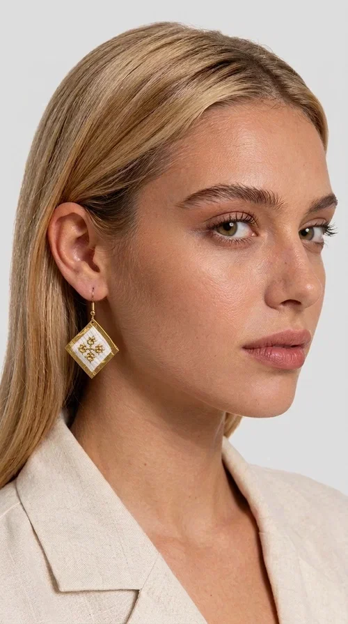 The embroidered statement earrings with geometric patterns in white and gold are the ideal choice for those seeking a piece of jewelry that combines traditional artistry with contemporary style. Designed by Greek Jewelry Designer Aikaterini Chalkiadaki, these handmade earrings represent a distinctive expression of Greek creativity and craftsmanship.

Their diamond shape, adorned with delicate embroidered decoration on white fabric and gold detailing, evokes the rich tradition of Greek folk art. The combination of brass and fabric creates a piece of jewelry that is light to wear yet striking in presence — perfect for elevating any look, from casual outfits to formal occasions such as weddings or christenings.

These dangling earrings pair perfectly with linen and minimal clothing, adding a boho-ethnic touch of elegance. They also make an excellent gift choice for women who appreciate unique, handmade jewelry with a Greek identity.