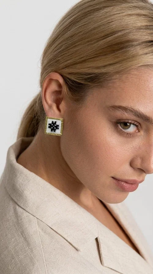 Embroidered statement earrings with geometric patterns are the ideal choice for those seeking an accessory that combines folk tradition with contemporary style. Designed by Greek Jewelry Designer Aikaterini Chalkiadaki, they are handmade jewelry pieces that highlight the charm of folklore aesthetics in a striking and timeless way.

Each pair features a square shape with a gold brass frame and finely worked embroidery in white and black, forming a geometric snowflake-inspired motif drawn from traditional Greek art. The result is a stud earring that immediately catches the eye, without being overly heavy or bulky — ideal for both everyday and formal wear.

They pair perfectly with casual looks and linen outfits, as well as with formal appearances at weddings, christenings, or summer events. The simplicity of their design makes them versatile, while the embroidery gives them a unique, artisan character.