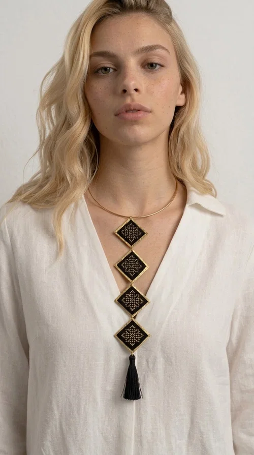 A statement embroidered necklace that combines tradition with modern aesthetics — ideal for every woman who wants to express herself through her jewelry. Designed by Greek Jewelry Designer Aikaterini Chalkiadaki, this striking long necklace features four diamond-shaped pieces in black fabric with gold-tone geometric embroidered designs, inspired by Greek folk tradition. It is completed with an elegant black tassel at the bottom, while the gold-tone choker base ring adds structure and style.

The necklace is made of brass and fabric, materials that ensure both durability and lightness. The handmade embroidered geometric motifs give each piece a unique character, while the combination of black and gold makes it versatile and timeless. It pairs equally well with casual and formal outfits, instantly offering a bold result.