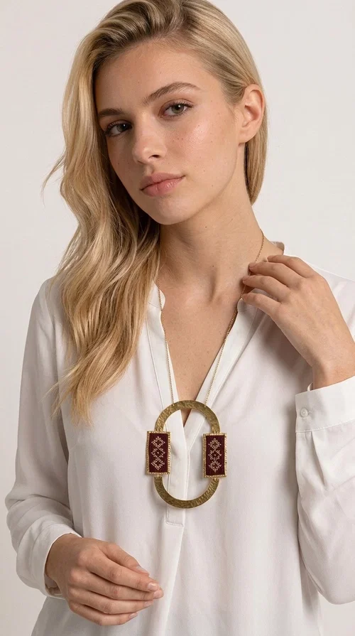 A statement embroidered necklace that combines folk tradition with modern aesthetics, creating a jewelry piece with a strong presence and character. Designed by Greek Jewelry Designer Aikaterini Chalkiadaki, this long necklace is an ideal choice for every woman who wants to express herself through fashion with boldness and style.

The necklace features a large oval element made of hammered brass in a gold tone, framed by two rectangular embroidered plates in deep burgundy color with geometric motifs inspired by Greek folk art and the tradition of embroidery. These details give the piece a folklore aura that suits both casual and formal outfits equally well.

The pendant necklace hangs from a delicate gold chain, creating a unique combination of handmade artistry and contemporary design. An ideal gift for friends, mothers, or yourself.