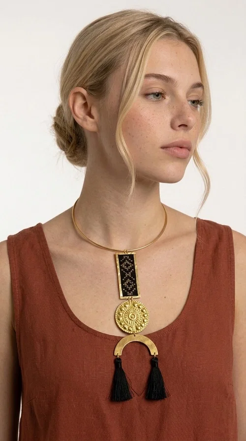 The statement embroidered necklace with geometric patterns in black is a jewel that never goes unnoticed — bold, handmade, and full of character. Designed by Greek Jewelry Designer Aikaterini Chalkiadaki, it combines the traditional art of embroidery with a modern aesthetic, creating a piece with strong personality and Greek soul.

The design features a gold-tone brass torque collar, from which hangs a rectangular embroidered panel with black geometric motifs inspired by Greek folk tradition. This is followed by a striking embossed brass disc with a sun motif, and at the bottom, a semicircular element adorned with three black tassels, adding movement and femininity to the overall look.

Ideal for looks you want to stand out in — from casual outfits with a linen dress or a plain t-shirt to evening appearances. Black and gold blend harmoniously with almost any clothing color.