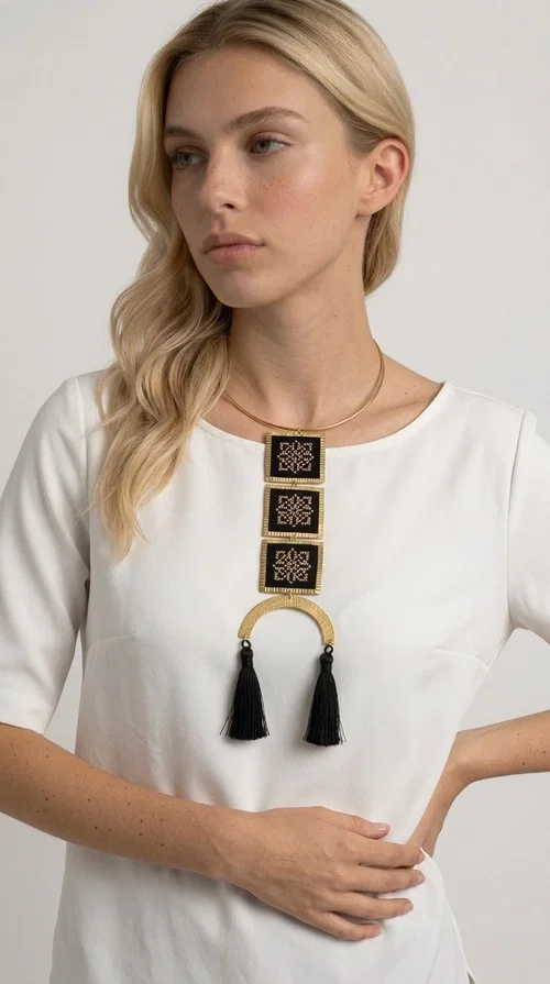 A statement embroidered necklace that combines the traditional art of embroidery with a modern, striking aesthetic — ideal for those who love jewelry with character and history.

Designed by Greek Jewelry Designer Aikaterini Chalkiadaki, this necklace features three square embroidered elements with geometric patterns in black and gold, inspired by Greek folk tradition. The base of the necklace is a round, snug brass choker in gold tone, from which hangs a series of embroidered squares ending in a semicircular brass element with two striking black tassels. The result is a long, impressive necklace with a strong presence that elevates any outfit.

The geometric patterns of the embroidery evoke a folklore aesthetic, while the combination of fabric and brass gives the piece a unique texture and weight. Ideal for a white or monochrome outfit you want to highlight.