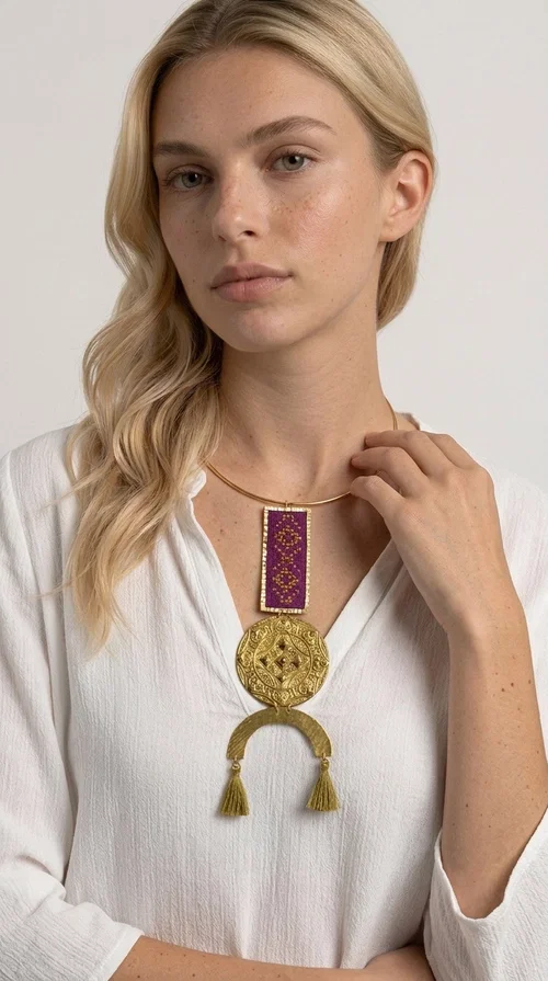 This statement embroidered necklace combines tradition with modern aesthetics, creating a piece of jewelry that instantly draws attention. Designed by Greek Jewelry Designer Aikaterini Chalkiadaki, it is a handmade creation that exudes a strong folklore character with clever contemporary styling.

The necklace features a rectangular embroidered panel in deep burgundy with geometric patterns, connected to an impressive golden circular brass disc with embossed motifs. The composition is completed with a semicircular element adorned with golden tassels, adding movement and vibrancy. It comes with a slim gold rigid collar that fits comfortably around the neck.

Ideal for elevating a simple white shirt, a boho dress, or even a stylish evening outfit. It instantly adds character and personality to your look.