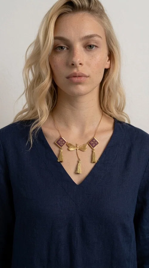 A statement embroidered necklace that combines the traditional art of embroidery with modern aesthetics — ideal for every woman who wants to express herself with style and originality. Designed by Greek Jewelry Designer Aikaterini Chalkiadaki, this handmade necklace is a true piece of wearable art, inspired by folk heritage and the geometric motifs of Greek tradition.

The necklace features two embroidered square elements in deep burgundy color with gold geometric patterns, joined by a central gold butterfly-shaped motif made of brass. Three gold tassels hang lightly, adding movement and charm to the whole. The gold-tone brass chain completes the elegant, striking look.

It pairs perfectly over simple, solid-colored outfits — such as a blue v-neck top — instantly adding character to your look. Suitable for daytime appearances, nights out, and as a unique gift for women who love striking, feminine jewelry.
