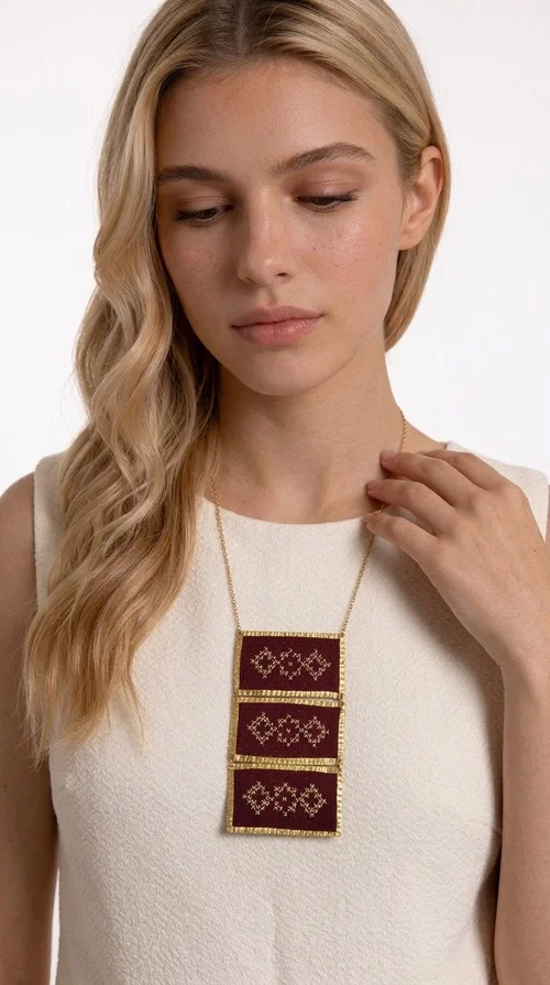 The statement embroidered necklace in burgundy with geometric patterns is a piece of jewelry that combines the traditional art of embroidery with a modern aesthetic, creating a result that immediately catches the eye. Designed by Greek Jewelry Designer Aikaterini Chalkiadaki, this striking necklace draws inspiration from folk tradition and the geometric motifs that characterize Greek handicraft. The deep burgundy color of the fabric, combined with gold brass, creates a rich, warm palette that suits every skin tone.

The necklace consists of three rectangular embroidered sections, placed one below the other and framed with gold brass featuring a textured finish. Each section showcases handmade embroidery with repeating geometric patterns in a folklore style, reminiscent of traditional Greek embroidery. The result is a long necklace with distinctive character, ideal for elevating a simple outfit or completing a boho-chic look.