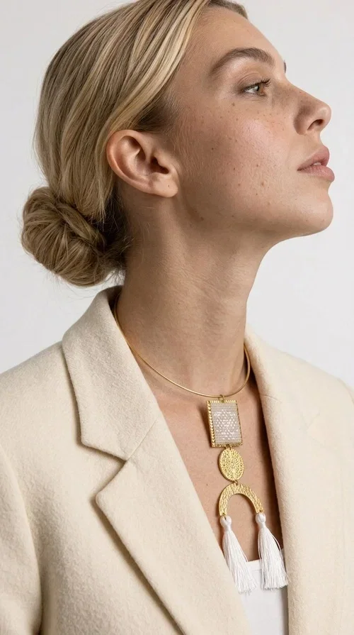 A statement embroidered necklace that combines folk aesthetics with modern elegance — the ideal piece for those who love style with character.

Designed by Greek Jewelry Designer Aikaterini Chalkiadaki, this handmade necklace consists of three main elements: a rectangular embroidered pendant with geometric patterns in cream tones, a gold circular disc with embossed decoration, and a semicircular gold base adorned with white fabric fringes. The necklace is worn on a gold stainless steel omega chain that fits comfortably around the neck, adding balance to the striking pendant.

The materials used — brass and fabric — create a unique result that resembles a work of art. The geometric embroidered patterns are reminiscent of traditional Greek techniques, while the overall composition has a distinctly contemporary character. Ideal for casual chic looks as well as evening outings, it pairs perfectly with a white t-shirt, an oversized jacket, or a simple dress.