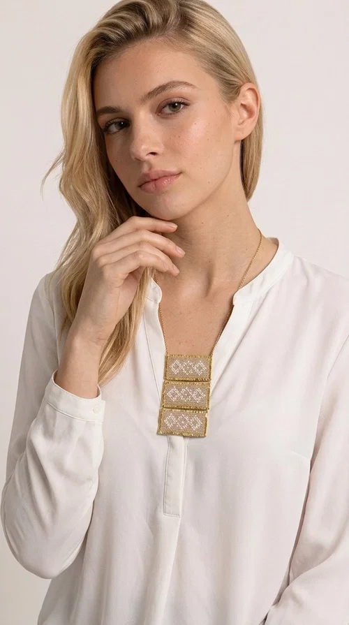The statement embroidered ivory necklace is a handmade creation that combines the traditional art of embroidery with a contemporary jewelry aesthetic, adding a unique, eye-catching touch to any outfit.

Designed by Greek Jewelry Designer Aikaterini Chalkiadaki, this necklace features three rectangular embroidered sections with ivory geometric motifs and gold details set in a brass frame. It hangs from a delicate gold chain that lends elegance and lightness, while the folklore-style geometric patterns give the piece a distinctive, ethnographic character. The combination of brass and fabric creates an intriguing contrast of materials that draws the eye.

Ideal for both day and evening wear, it pairs beautifully with white or monochrome outfits to highlight its geometric patterns. The ivory color makes it versatile and timeless, perfect for summer looks as well as formal or business occasions.