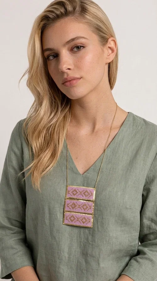 An embroidered necklace that combines tradition with contemporary aesthetics — the Statement Embroidered Necklace in Lilac with Geometric Patterns is the jewel that turns any look into a style statement. Designed by Greek Jewelry Designer Aikaterini Chalkiadaki, this long necklace carries the distinctive energy of folklore aesthetics in a bold, striking version.

Three rectangular brass plates in a gold hue are covered with handmade embroidery in lilac and gold tones, featuring repeating geometric motifs inspired by Greek tradition. They hang from a delicate gold chain, creating a long necklace with a statement presence that suits both casual and elegant looks.

Ideal to pair with linen, cotton or evening wear, this necklace adds character to any outfit — from summer outings to formal occasions. The lilac hue is romantic and versatile, while the geometric patterns bring a strong personality.