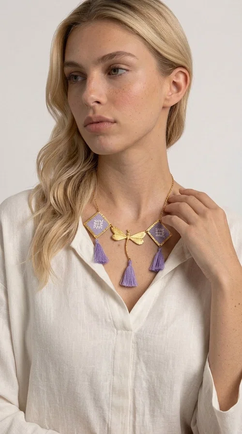 A statement embroidered necklace that blends the traditional art of embroidery with a contemporary aesthetic — ideal for every woman who wants to express her uniqueness with style.

Designed by Greek Jewelry Designer Aikaterini Chalkiadaki, this striking necklace features two diamond-shaped pendants with handmade embroidery in shades of lilac, a gold brass facing deer (libelle) charm at the center, and four fabric tassels in lilac tones that move gracefully with every step. The gold brass chain that fastens at the back of the neck gives the piece a delicate, elegant silhouette. The geometric embroidery patterns are reminiscent of authentic Greek folk motifs, evoking the rich tradition of Greek handicraft.

Perfect for summer looks, celebrations, or everyday moments that call for something special, this handmade necklace pairs beautifully with a white shirt, a dress, or a boho outfit. Pair it with simple earrings to let it stand out to the fullest.
