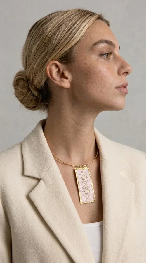 The embroidered pink necklace with geometric patterns is a statement piece that combines the traditional art of embroidery with a modern, striking result. Designed by Greek Jewelry Designer Aikaterini Chalkiadaki, this unique necklace draws inspiration from folk tradition and the folk aesthetic that defines the Folklore Jewelry collection.

The embroidered rectangular pendant, in a soft pink shade, is adorned with gold geometric motifs reminiscent of traditional embroidery, while the gold brass metal frame gives it structure and elegance. It is worn on a delicate gold torque-style neck hoop that fits comfortably and instantly elevates any outfit, from casual to formal.

Ideal for women who love handmade jewelry with character, it is the perfect choice as a gift or to refresh your own jewelry collection with a piece full of soul.