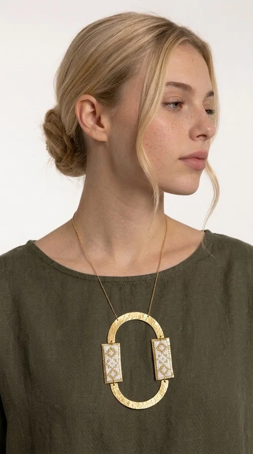 An embroidered necklace that blends tradition with modern design — the Statement necklace KOL-4745 is the ideal choice for every woman who wants to express herself with style and authenticity.

Designed by Greek Jewelry Designer Aikaterini Chalkiadaki, this striking long necklace features a handmade oval brass element in a gold tone, framed by two rectangular embroidered plaques made of white fabric with geometric patterns inspired by Greek folk tradition. The gold-tone chain adds an elegant, modern touch, while the embroidered motifs bring a warm, handcrafted feel.

Perfect for pairing with simple outfits in neutral shades — such as khaki, white or beige — it turns any look into a true statement. Suitable for both everyday wear and special evening occasions.