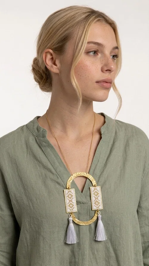 An embroidered necklace that combines tradition with contemporary design — the Statement Necklace KOL-4757 is the ideal choice for every woman who wants to express herself through her jewelry. Designed by Greek Jewelry Designer Aikaterini Chalkiadaki, this handmade necklace is a unique combination of fabric and brass, inspired by folk art and the geometric motifs of Greek tradition.

Two embroidered rectangular plates with geometric patterns in white and gold frame a hammered gold brass circle, while three light-colored tassels add movement and character. The long gold chain completes a composition that is both statement-making and harmonious. Ideal for summer looks, casual outfits, and evening moments when you want to stand out.