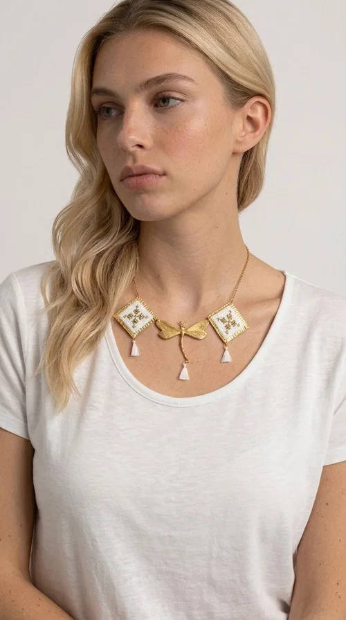 The statement embroidered necklace in white with geometric patterns is a piece of jewelry that combines the traditional art of embroidery with a modern, striking result. Designed by Greek Jewelry Designer Aikaterini Chalkiadaki, this unique necklace showcases Greek craftsmanship in all its glory.

The necklace features two embroidered square elements in white with gold geometric motifs inspired by Greek folk tradition, joined by a gold dragonfly-shaped element. Three delicate white tassels add movement and joy to the piece, while the gold chain completes the look with elegance. The contrast between the white fabric and the brass creates a result that is both delicate and striking.

Ideal for summer looks, brunch, weddings, or evening outings, this handmade necklace pairs equally well with a simple white T-shirt or a boho dress. It also makes an excellent gift choice for women who love unique, handmade jewelry.