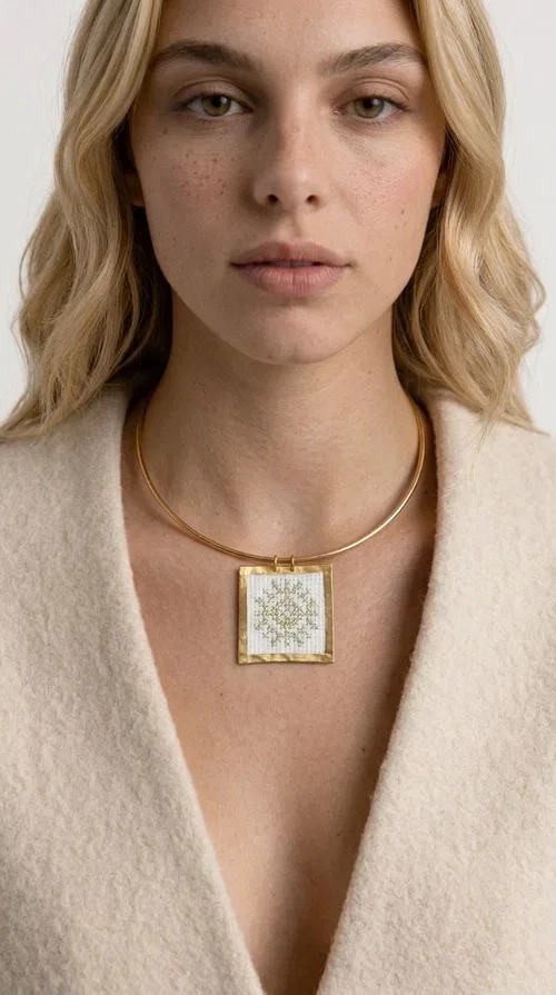 An embroidered necklace that combines tradition with modern aesthetics — the Statement Necklace KOL-4721 is a handmade creation that stands out for its refined detail and unique character. Designed by Greek Jewelry Designer Aikaterini Chalkiadaki, this piece pays tribute to the folk Greek art of embroidery, rendering geometric motifs on white fabric with gold details.

The rectangular embroidered pendant, featuring a handcrafted gold-tone brass frame, hangs from a classic gold omega-style necklace chain, which adds stability and elegance to the overall look. The folklore geometric patterns on the white fabric create an eye-catching contrast, while the overall aesthetic moves between ethnic and contemporary style.

Ideal for pairing with linen or wool clothing in neutral tones, it suits both casual and more formal looks. A true statement piece for the neck that needs no other additions to shine.